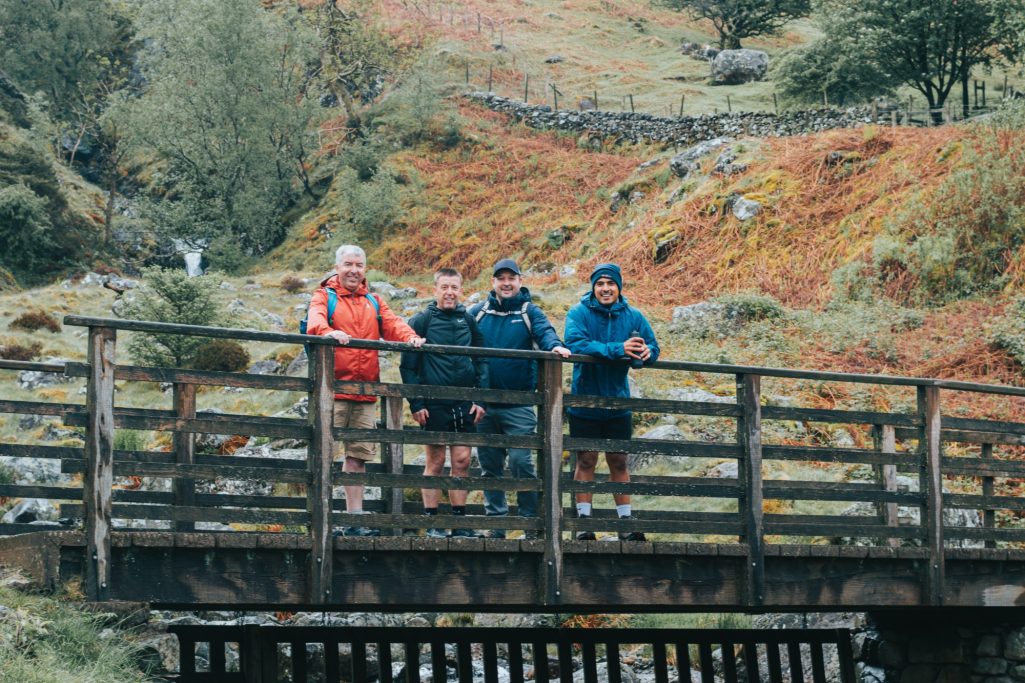 Four people on a wooden bridge in a wooded area with autumn foliage.