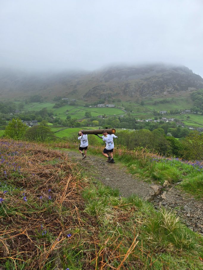 Two hikers on a path, carrying a log with lush green hills and fog in the background.