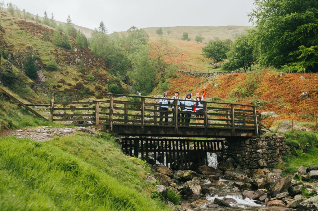 Wooden bridge over a stream, surrounded by green hills and trees on a rainy day.