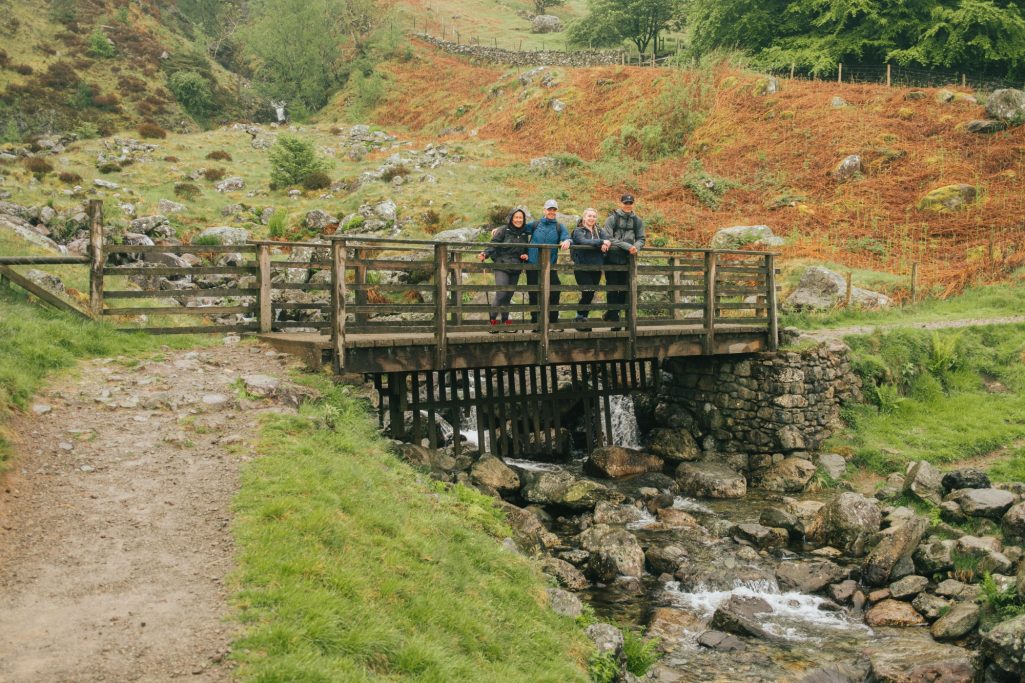 Three people standing on a wooden bridge over a stream in a rural landscape.