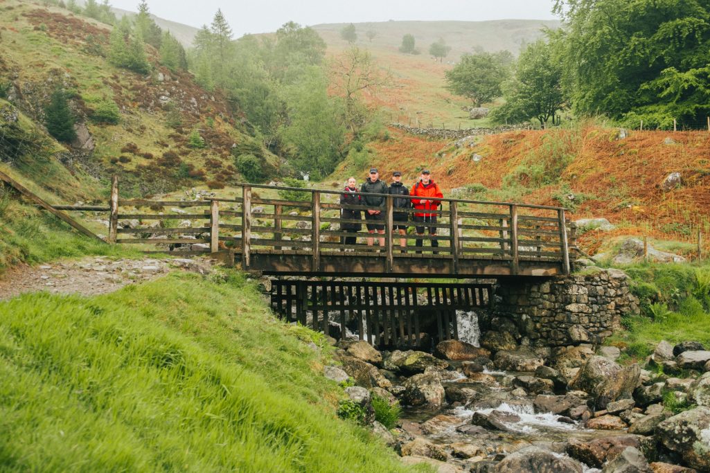 Three people standing on a wooden bridge over a stream in a lush green landscape.