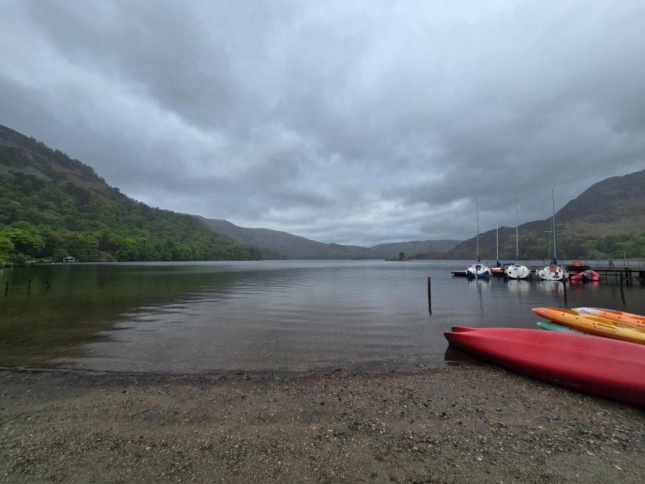 Calm lake scene with moored boats, grey skies, and surrounding green hills.