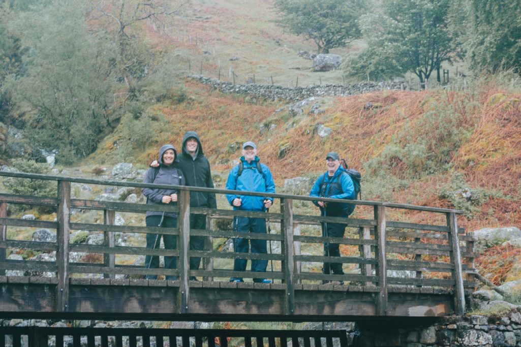 Group of four people standing on a wooden bridge in a rural, autumn landscape.
